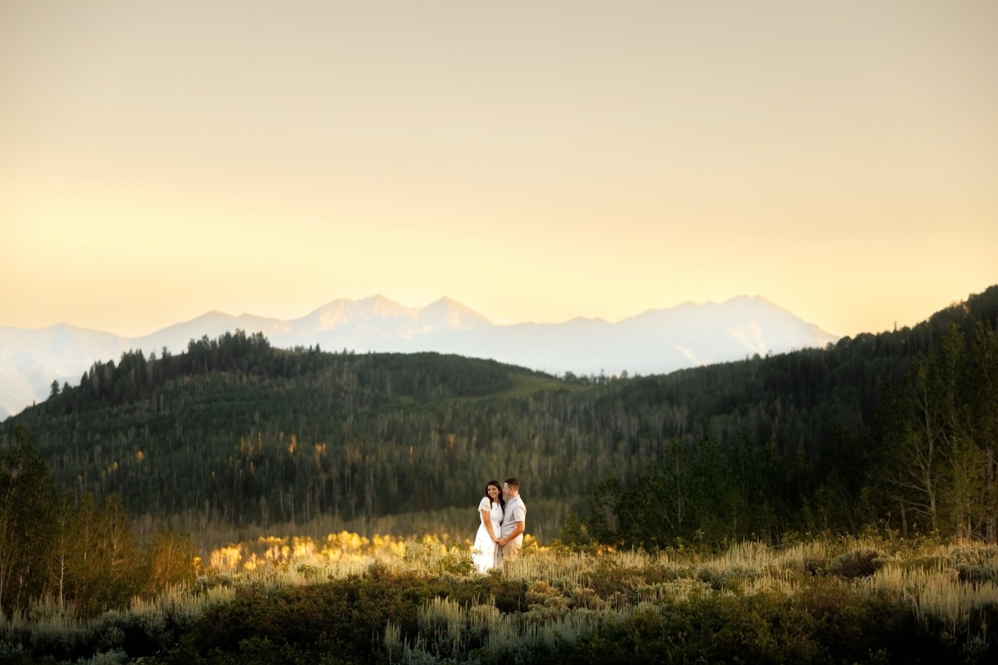 A couple standing small in a wide mountain valley at last light, ridgelines stacked in layers behind them