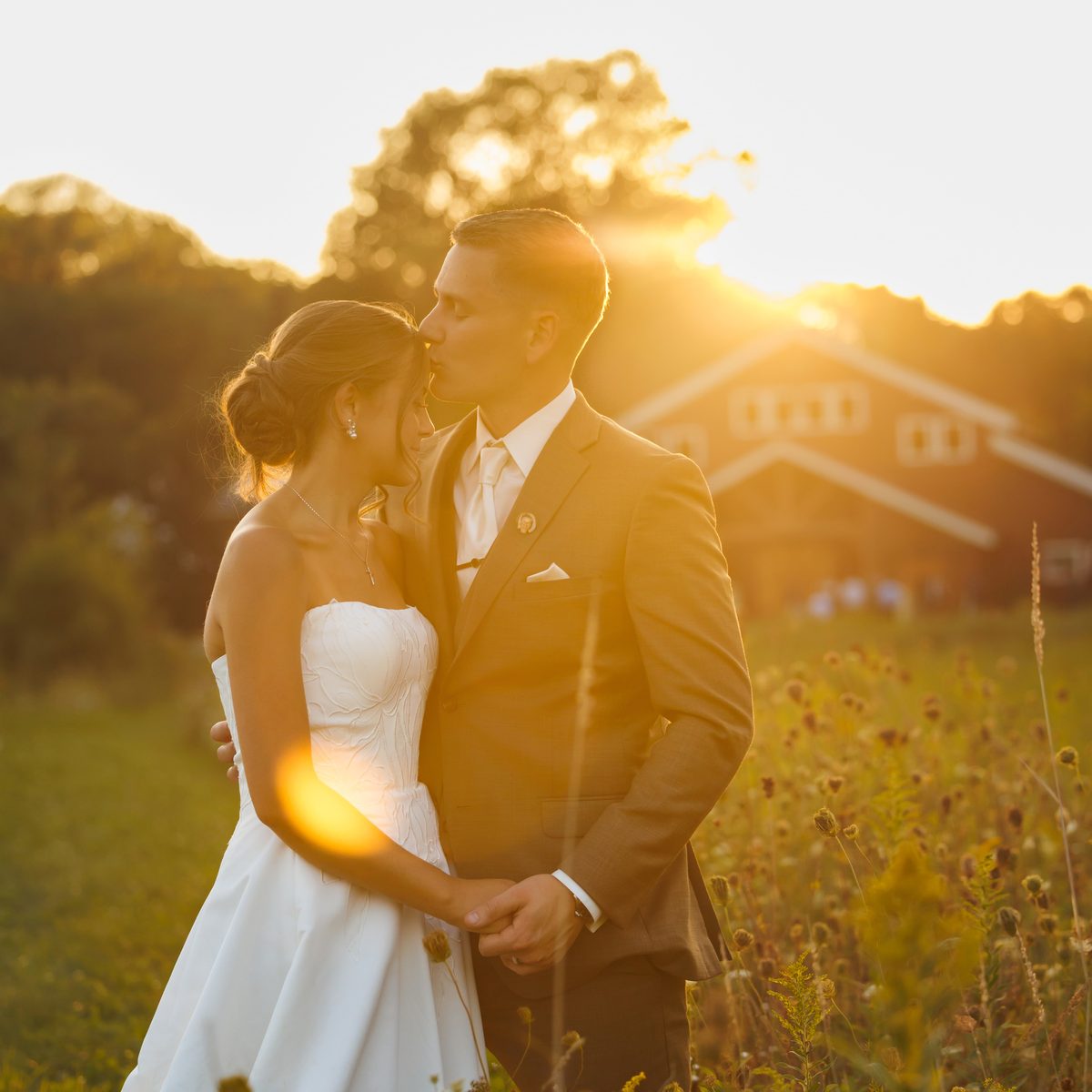 A couple holding each other in a wildflower field as the sun sets behind the barn